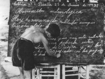 Un jove colon escriu a la pissarra mentre es desenvolupa la classe a l'aire lliure. DEL LLIBRE 
